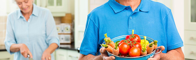 Senior Man Holding Vegetable Bowl With Woman Cutting Vegetables.